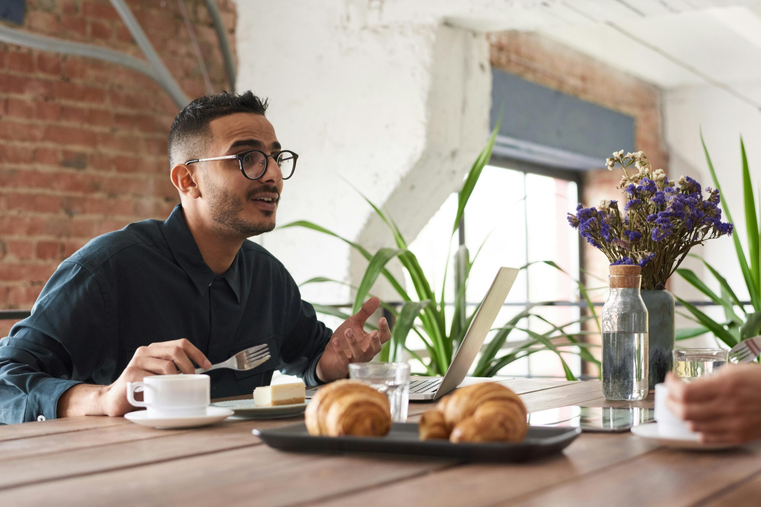 A man in glasses engaging in conversation over brunch with croissants and coffee.